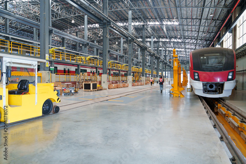 Red electric train undergoing inspection within expansive industrial warehouse with tracks and machinery; technician wearing red uniform observes vehicle condition during maintenance.