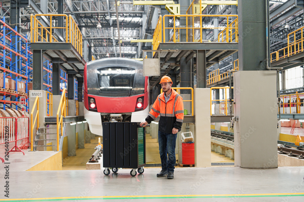 Obraz premium Skilled maintenance worker pushes wheeled cart filled with tools towards large electric train within spacious railway maintenance hall for servicing and repairs.