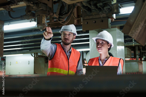 Serious technician and engineer intently inspect complex industrial machinery during equipment evaluation within bright, modern factory setting focusing on detailed maintenance work.