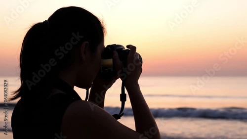 Silhouette of female travel photographer using digital dslr camera on tropical beach during vibrant orange golden hour sunrise