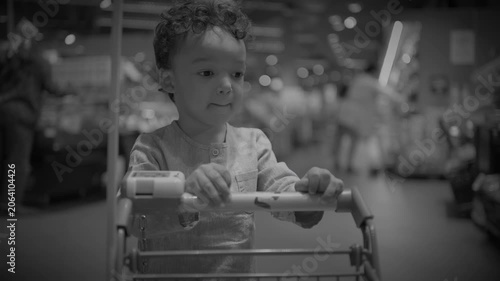 Young child standing in a shopping cart in supermarket aisle, holding handle and looking right