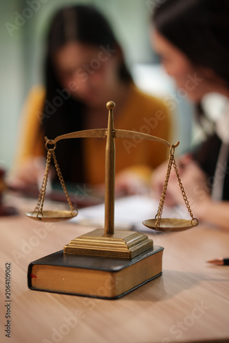 Golden brass scales of justice standing on law book with lawyers discussing case in blurred background.