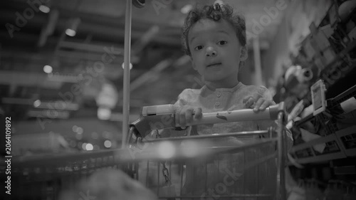 Curly-haired toddler reaching from shopping cart in supermarket aisle