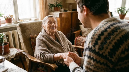 Young man holding hands with his elderly grandmother and talking. Grandson visiting senior woman at home. Family connection and care concept