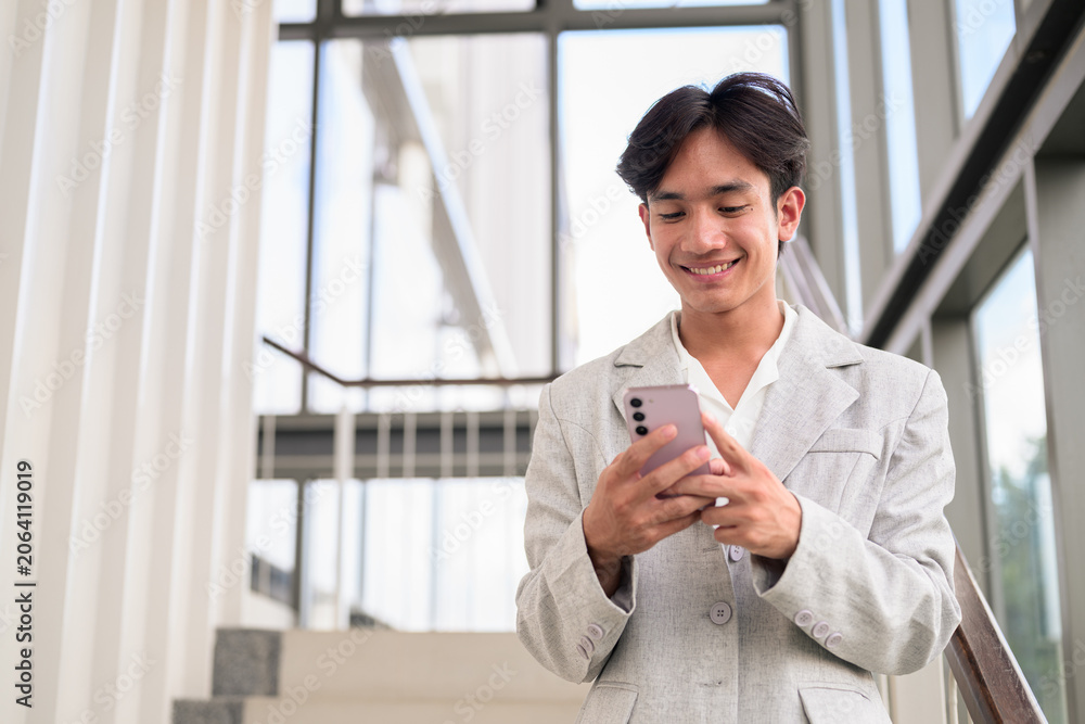 Fototapeta premium Smiling young nonbinary Thai business professional texting on smartphone on office stairs