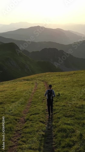Young man with a backpack hiking on a scenic mountain path. Beautiful golden hour light illuminates the lush green hills and distant peaks