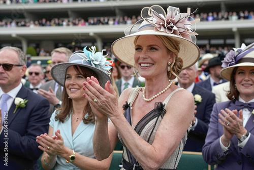 A woman in elegant dress and fancy decorative hat, clapping and cheering with other elegantly dressed spectators watching horse racing in a crowded racetrack. High society, glamour socializing event.