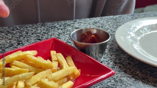 Hand holding french fry over red bowl with ketchup