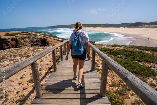 Visiting Algarve coast in Portugal. Beautiful middle-aged woman tourist walking down wooden stairs to sandy Bordeira beach and enjoying vacations over ocean on summer day. Back view