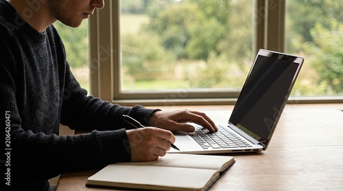 Young man working on laptop and writing in notebook by window.