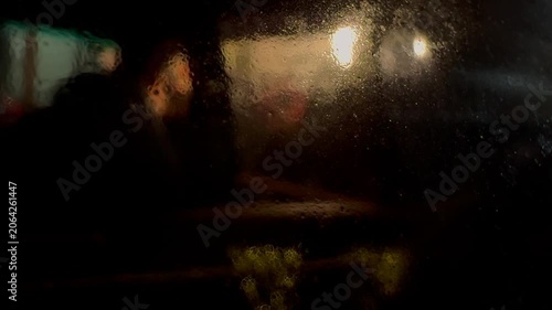 Blurred silhouettes of people moving through a city street at night, viewed through a car window, creating an atmospheric urban scene with motion, reflections, and low-light ambiance