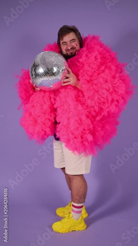 Vertical 4K video. 60 fps. Young adult white man with beard, stickers on male face, pink tulle fur coat, yellow sneakers, shorts, striped socks holds silver glitter disco ball on lilac. Diverse beauty