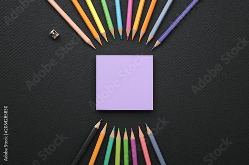 Light purple sticky note surrounded by arranged colored pencils on dark slate surface with stationery supplies creative arrangement background education planning composition business contrast