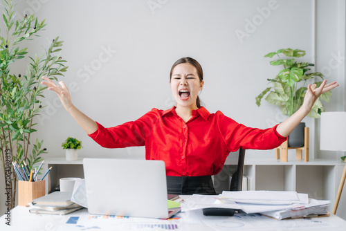Fotografie Overwhelmed Employee Exasperation: A young woman in business attire throws her hands up in exasperation, her face a mask of frustration and stress, while seated at a cluttered desk with a laptop