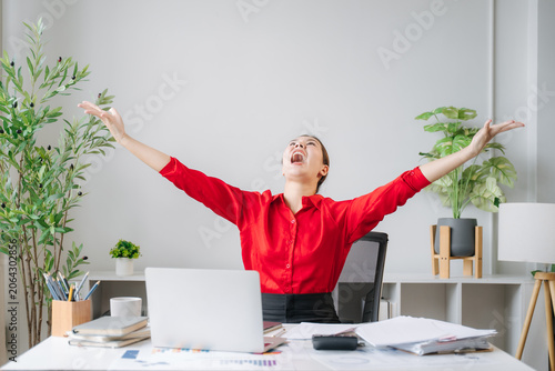 Bilde på lerret Triumphant Entrepreneur: A woman in a bright red shirt throws her arms wide open in a moment of pure elation and success, seated at her desk surrounded by office supplies and plants