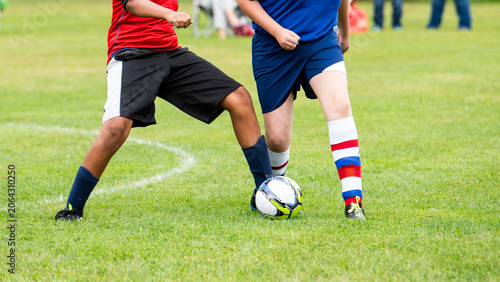 Fotografija Two youth soccer players battle for the ball on a grass field, legs and striped socks in motion as a white ball sits between red and blue uniforms, faces out of frame for release free use