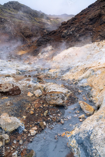 vertical photograph of rocks, mud, steam and boiling water in Seltun, a geothermal area in Reykjanes Peninsula in Iceland on a spring day