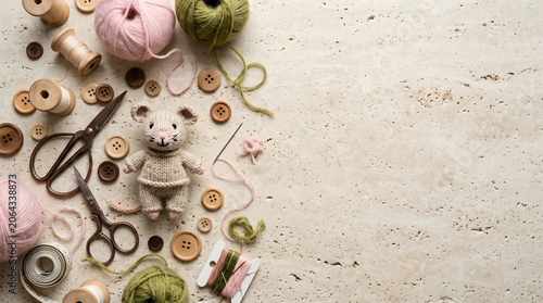 Cozy handmade craft composition on textured stone background with knitted mouse toy, wooden buttons, vintage scissors, pink and olive green wool yarn, and tools with large copy space.