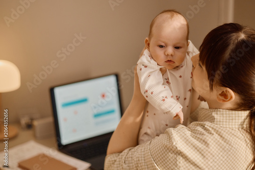 Caucasian mother holding baby at home near laptop, balancing infant care with online tasks, showing modern parenting routine. Ideal for maternity, childcare, work life balance marketing