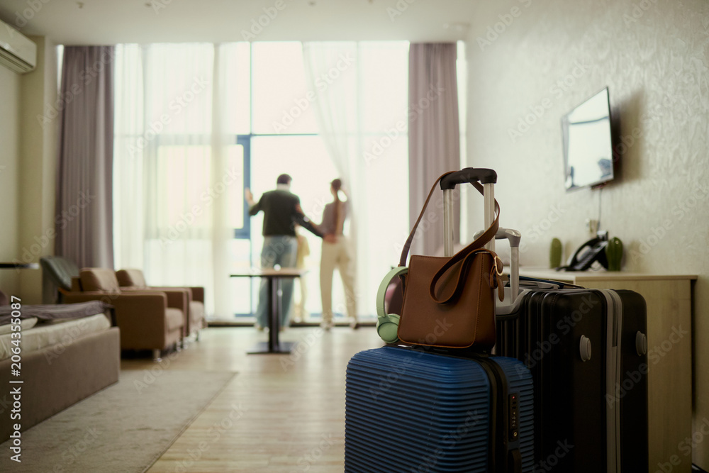 Obraz premium Wide angle view of young couple arriving in hotel room, unpacking luggage and starting vacation stay, checking view from window. Travel booking, hospitality marketing, family tourism promotion