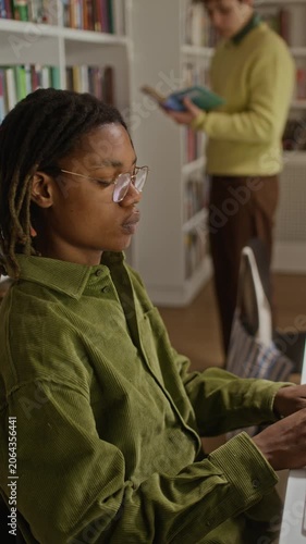 Vertical side view shot of African American teenage boy with glasses typing on computer in library while Caucasian teenage boy reading book in background. Video perfect for educational marketing