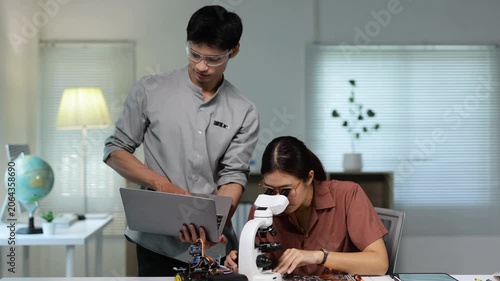 Two young asian engineers collaborating on a robotics project in a technology lab, with the woman looking through a microscope at a circuit board and the man assisting her with a laptop