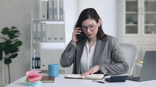Frustrated female executive gesturing and arguing during a difficult phone call while flipping through her notebook at her modern office desk, feeling angry and under pressure