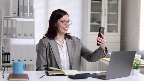 Smiling professional woman in a suit having a pleasant business video conference on her mobile phone while sitting at her desk in a modern office with a laptop in the background