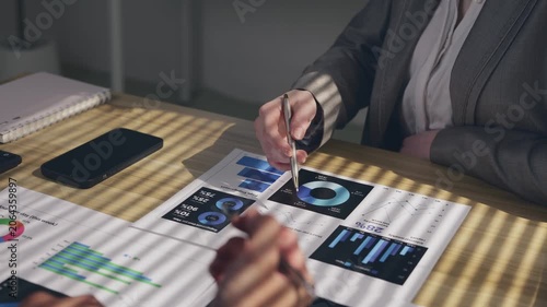 Two business professionals sitting at a wooden desk discussing and analyzing financial performance data with charts and graphs, gesturing with pens during a corporate strategy meeting