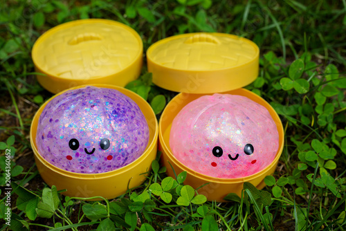  Two glitter slime dumpling toys with kawaii faces in purple and pink sitting in open yellow steamer containers on green grass