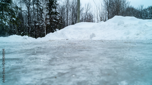 Fotografie A dramatic low-angle ground-level photograph looking across a thick, textured sheet of frozen glare ice toward large plowed snowbanks and winter forest trees on a cold, overcast day