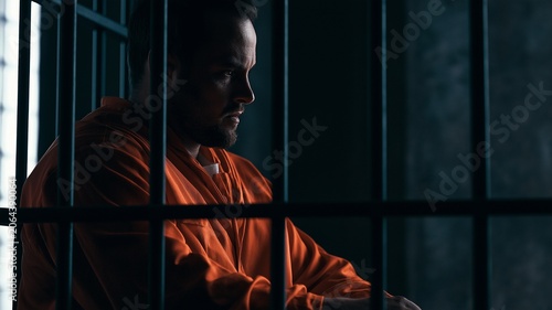 A solitary inmate in an orange prison uniform sits behind metal bars inside a correctional facility. symbolizes incarceration, criminal justice, punishment, confinement
