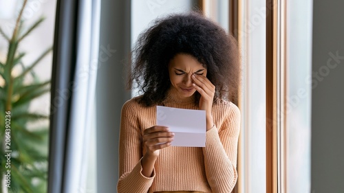 A young woman stands by a window reading a letter or document while wiping away tears. emotions of sadness, grief, disappointment