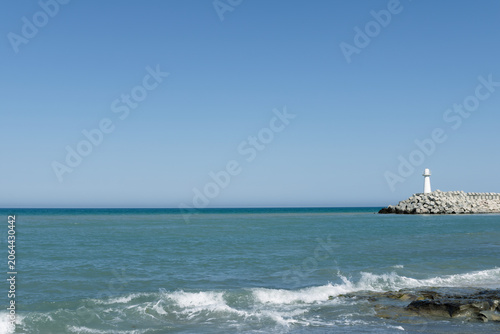 White lighthouse on rocky breakwater with turquoise sea and waves in foreground, clear blue sky, peaceful coastal seascape on sunny summer day.