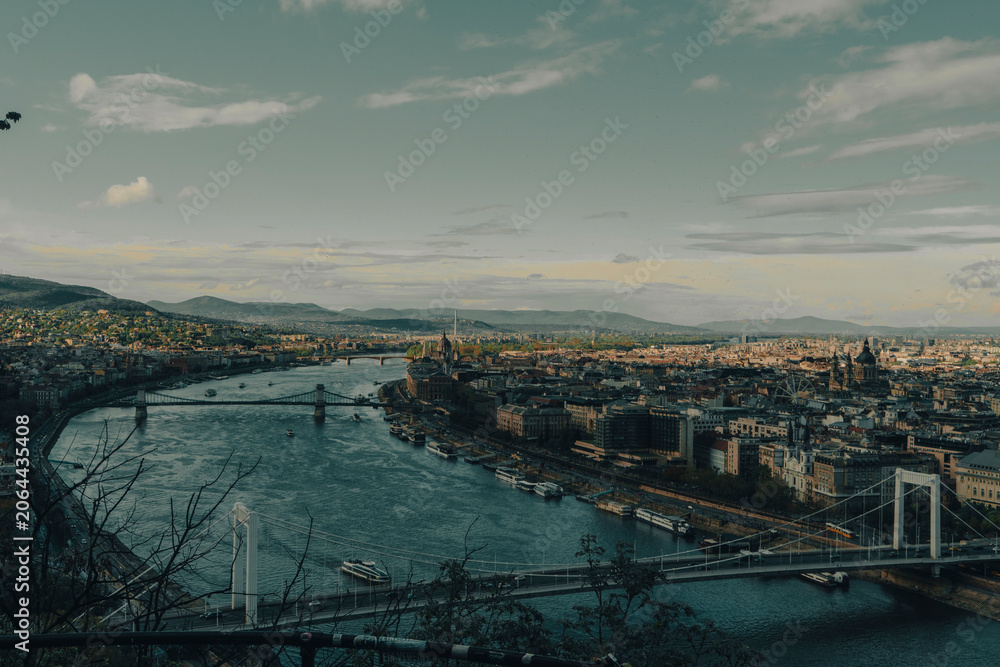 Fototapeta premium ​Horizontal panoramic high angle view of a city river landscape with multiple historic bridges. Grand urban cityscape and mountain skyline under a cloudy teal sky, shot in Budapest, Hungary.