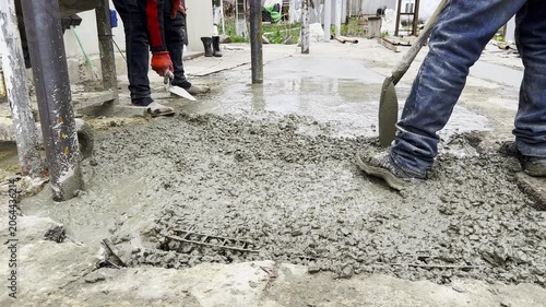 Male hands in protective gloves uses trowel to spread and level fresh cement in yard. Another worker distributes and moves the mixture with a shovel preparing the material during construction process