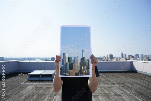 Person holding up picture of city buildings against clear sky over rooftop