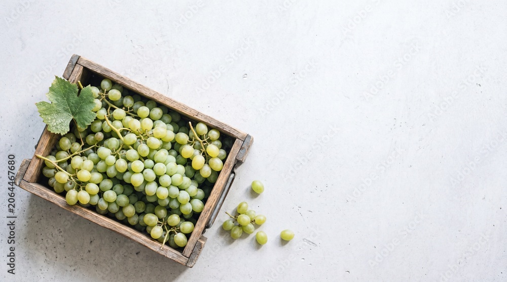 Fototapeta premium A wooden crate filled with fresh green grapes. Some grapes are scattered around the crate. The background is light and neutral, emphasizing the fruit.