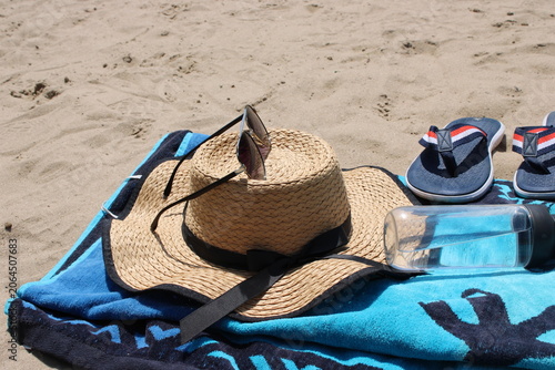 Straw sun hat with sunglasses resting on a blue beach towel alongside flip-flops and a water bottle on sandy beach surface under bright sunlight