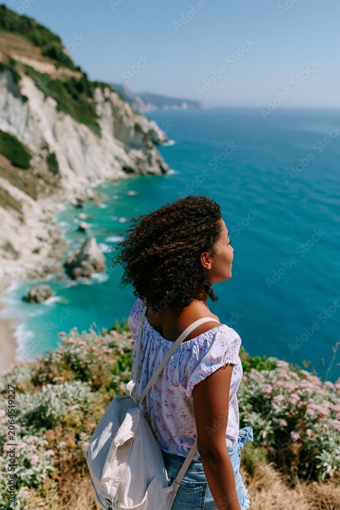 Fototapeta premium Young woman with curly hair wearing a floral top and backpack stands on a cliff overlooking the ocean with rocky coastline and blue sky in the background