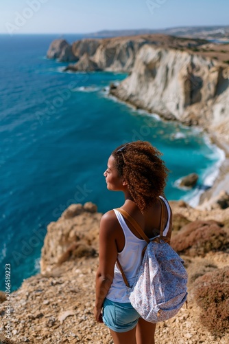 Young woman with backpack stands on cliff edge overlooking the ocean, rocky coastline and clear blue water visible in the background