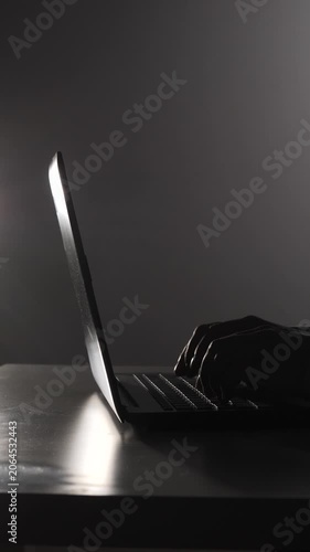 Vertical video person's hands typing on a laptop keyboard with a bright screen in a dimly lit setting, creating a silhouette, representing concepts of technology, hacking, and online security