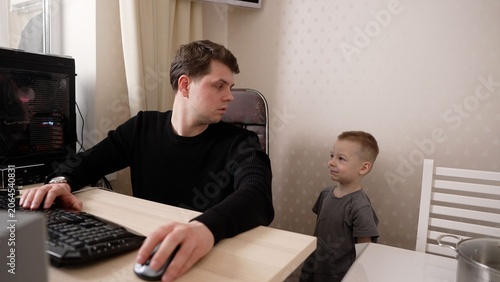 A young father tries to concentrate on his computer while his little son playfully distracts him. The boy seeks attention, showing the challenges of remote work and family life at home