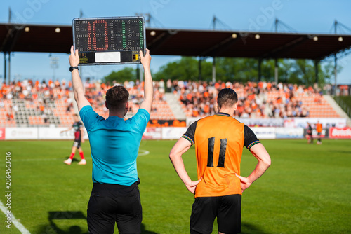 Foto A soccer referee holding an electronic substitution board next to a player in an orange jersey waiting to enter the pitch