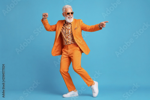 A man with gray hair and a beard dances joyfully in an orange suit. He wears sunglasses and white shoes. The background is blue. The scene shows a fun moment of celebration.