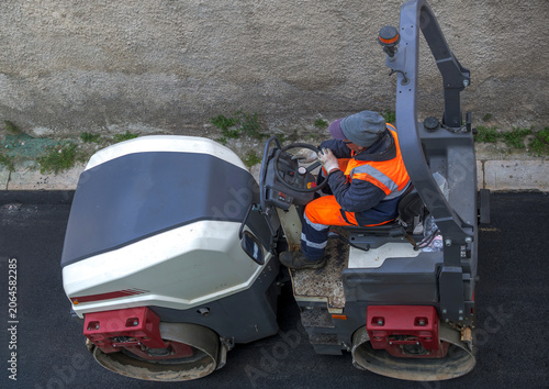 High angle view of a double drum vibratory roller compactor actively compacting fresh black asphalt during street construction, operated by an industrial worker in a safety vest.