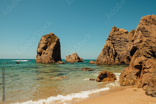 beach Ursa, Portugal. Coastal view with rock formations  enjoying the beach on a sunny day near the water.  The sun shines bright over the clear blue ocean on a warm day.