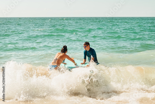 beach Ursa, Portugal.  Two friends sit on a surfboard while holding drinks as waves crash around them. Another person is seen paddling in background. sunny warm on beach.  waves crash  in warm weather