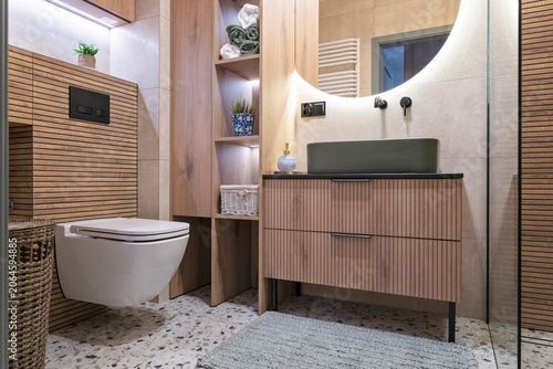 Modern bathroom interior with bathroom sink on wooden cabinet and toilet on tiled floor. Contemporary interior with luxurious mirror on the wall. Real estate.
