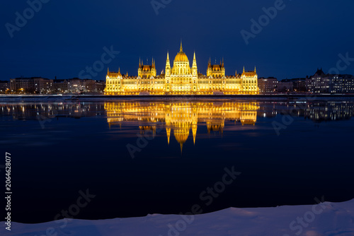 Winter night landscape of the illuminated Hungarian Parliament Building with reflection on the Danube River and snow in the foreground, Budapest